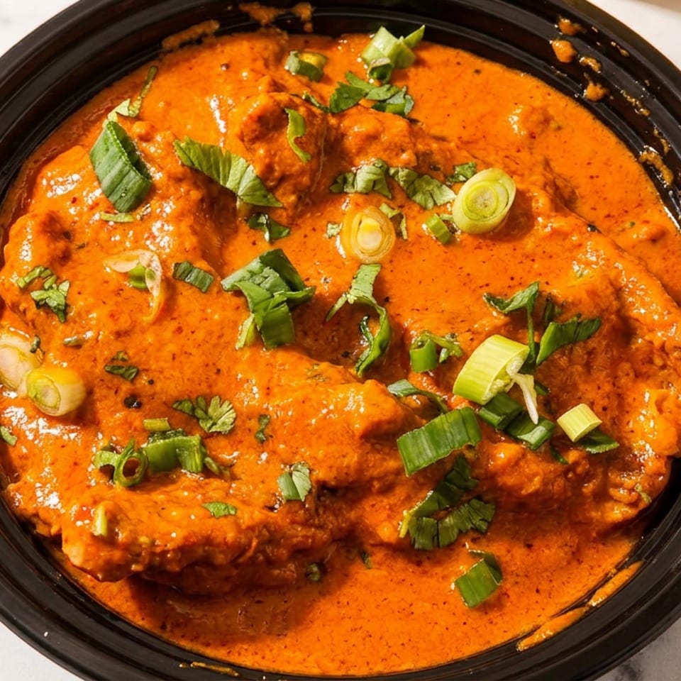A black bowl holds tender chicken in creamy orange curry sauce, garnished with chopped green onions and cilantro.